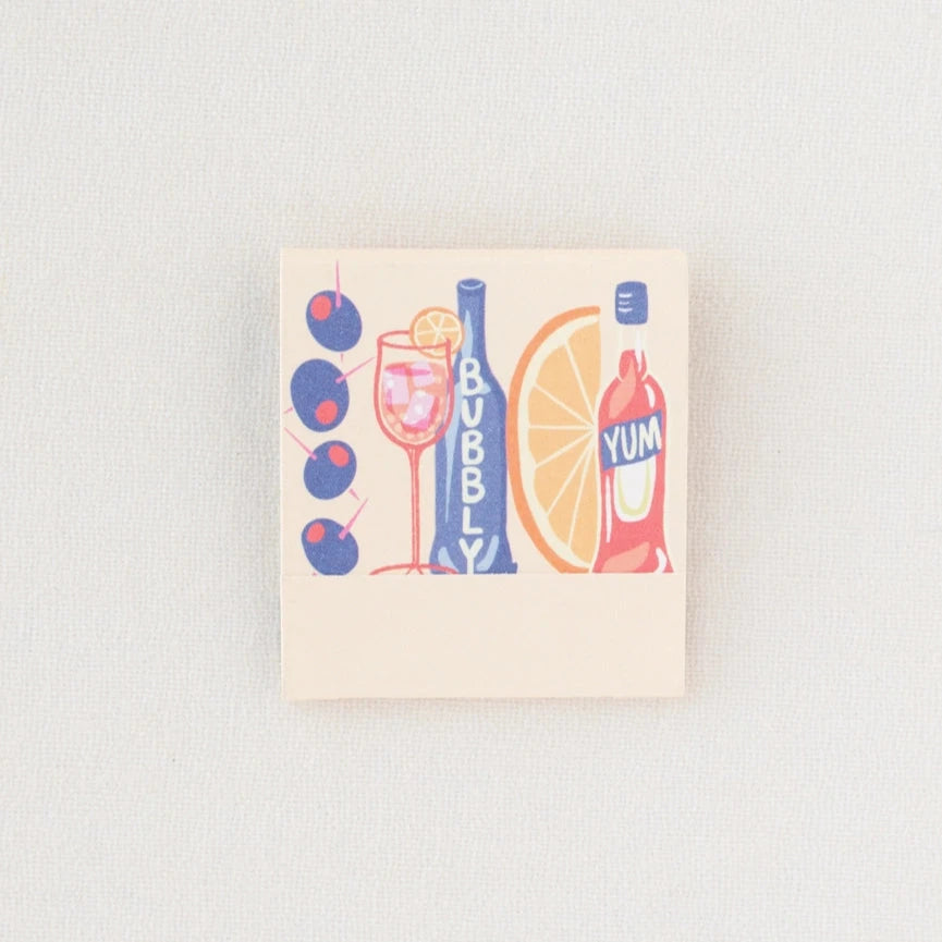 Spritz matchbook shown closed, with image on cover of Campari, orange wedge, bubbles, spritz in a glass and olives on a pick.
