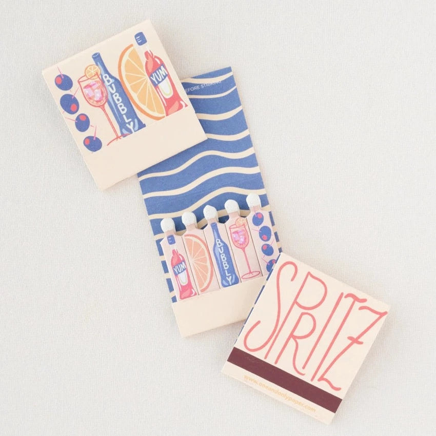 Spritz matchbook open to reveal that the matches are in the shape of Campari, orange wedge, bubbles, spritz in a glass and olives on a pick.
