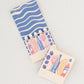 Spritz matchbook open to reveal that the matches are in the shape of Campari, orange wedge, bubbles, spritz in a glass and olives on a pick.