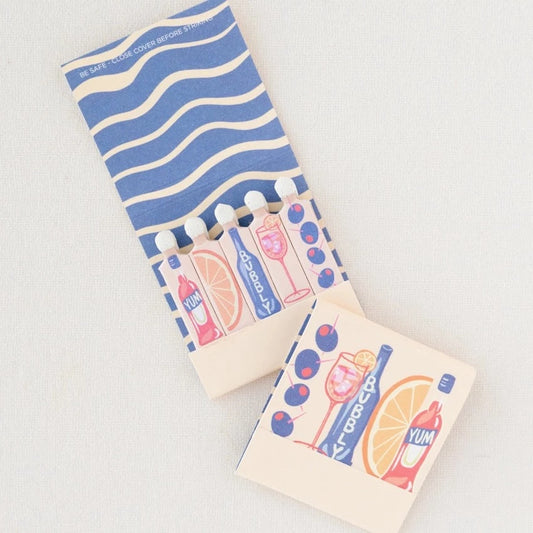 Spritz matchbook open to reveal that the matches are in the shape of Campari, orange wedge, bubbles, spritz in a glass and olives on a pick.