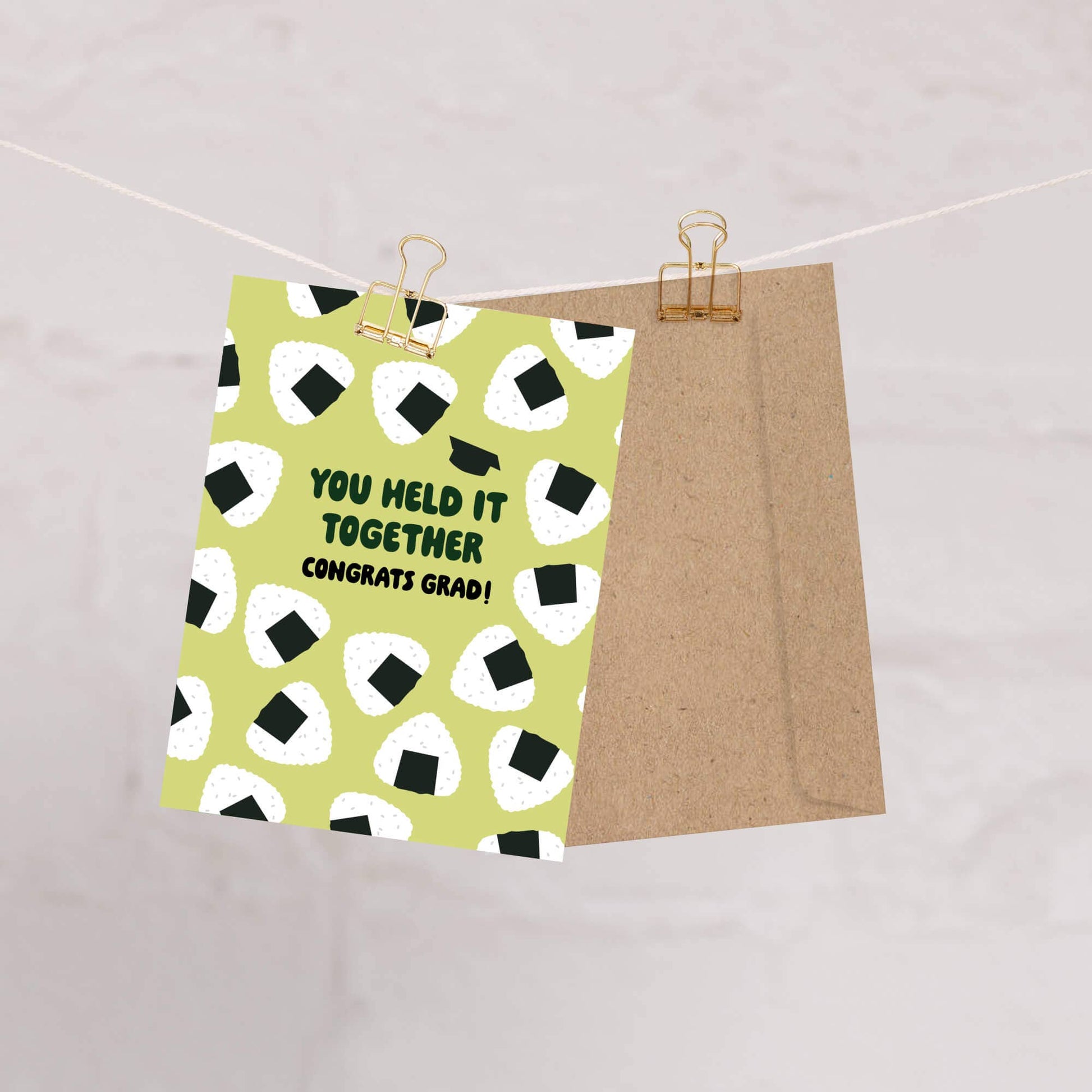 Graduation card with onigiri rice ball pattern on a green background, held up by a clothesline.