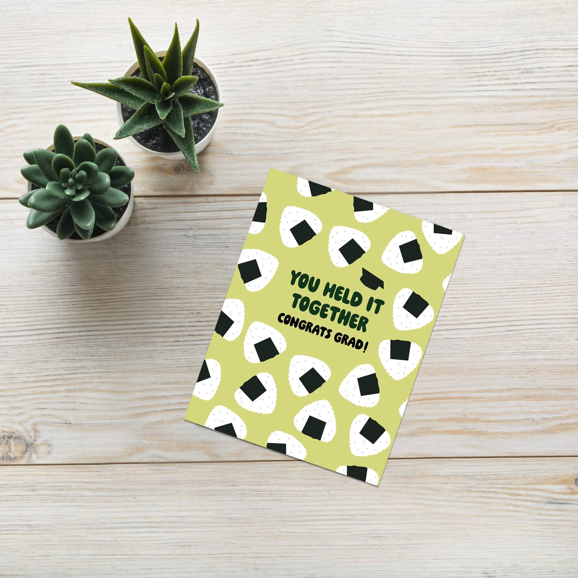 Graduation card with onigiri rice ball pattern on a wooden surface with small plants.