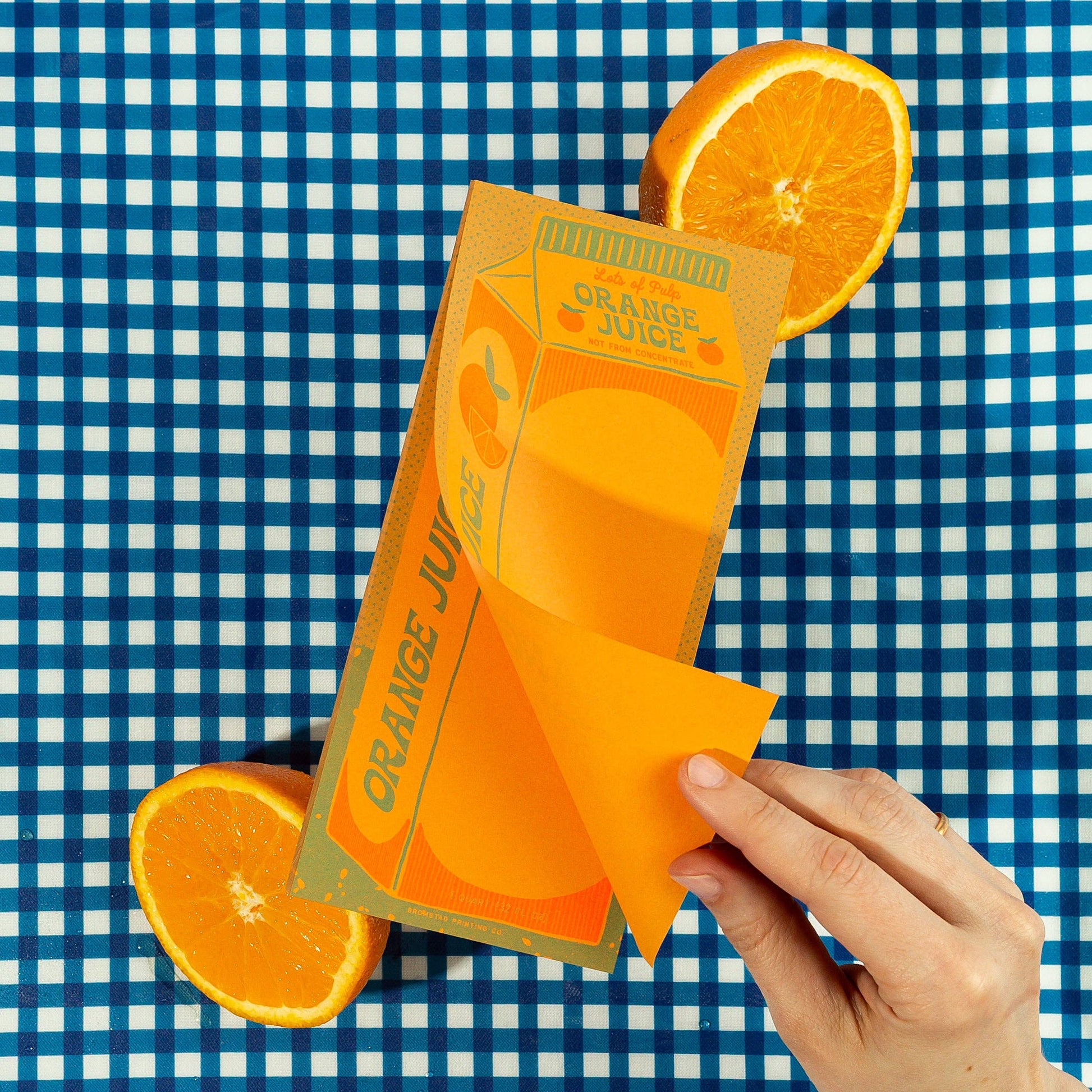 Carton of orange juice illustration on rectangular notepad pictured with orange halves on a blue and white checkered background 