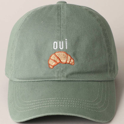 A green baseball cap with 'oui' written on it and an embroidered croissant below the text.