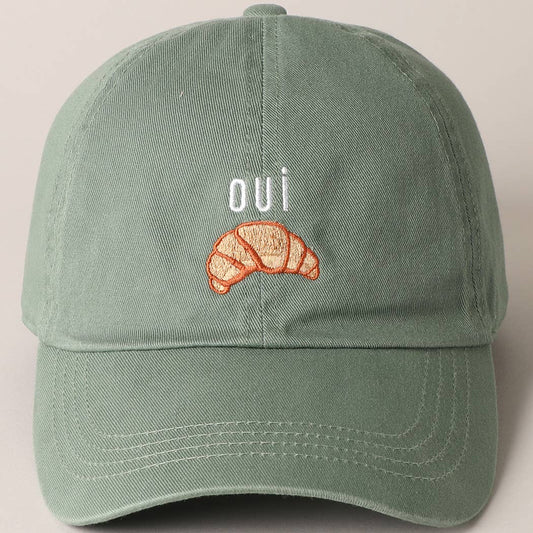 A green baseball cap with 'oui' written on it and an embroidered croissant below the text.