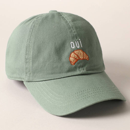 A green baseball cap with 'oui' written on it and an embroidered croissant below the text.