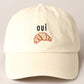 A cream baseball cap with 'oui' written on it and an embroidered croissant below the text.