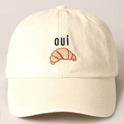 A cream baseball cap with 'oui' written on it and an embroidered croissant below the text.