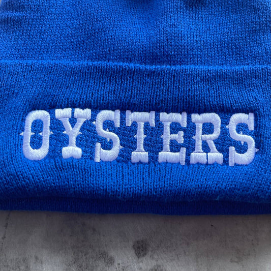 close up of the blue knit beanie with white embroidered text that reads "oysters" 