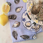 light blue tea towel with oysters and lemon design pictured on marble surface with lemon wedges and oyster shells 