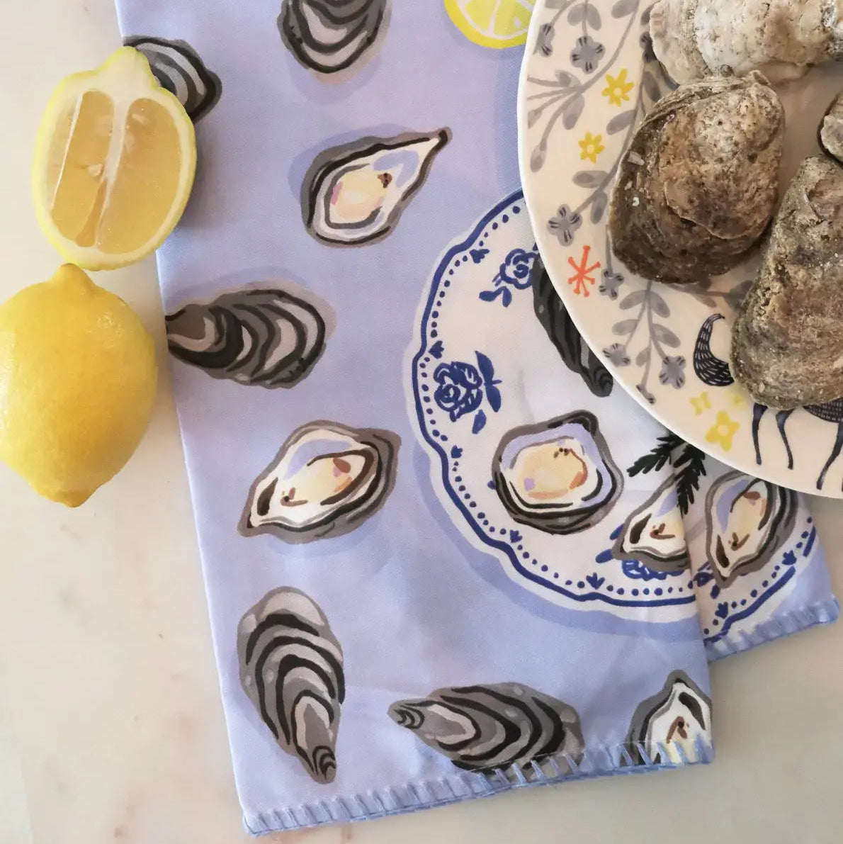 light blue tea towel with oysters and lemon design pictured on marble surface with lemon wedges and oyster shells 