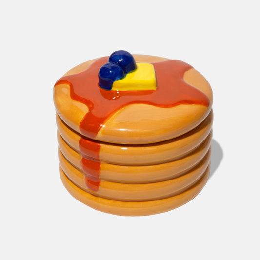 Ceramic storage jar made to look like a stack of pancakes with a decorative lid with a pad of butter and blueberries on a white background
