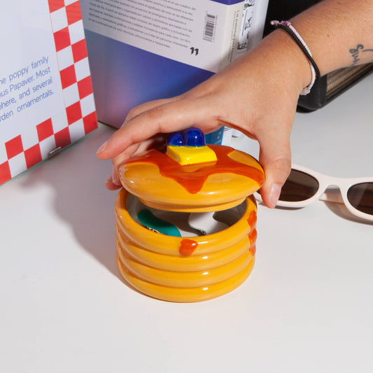 Ceramic storage jar made to look like a stack of pancakes with a decorative lid with a pad of butter and blueberries on a white background