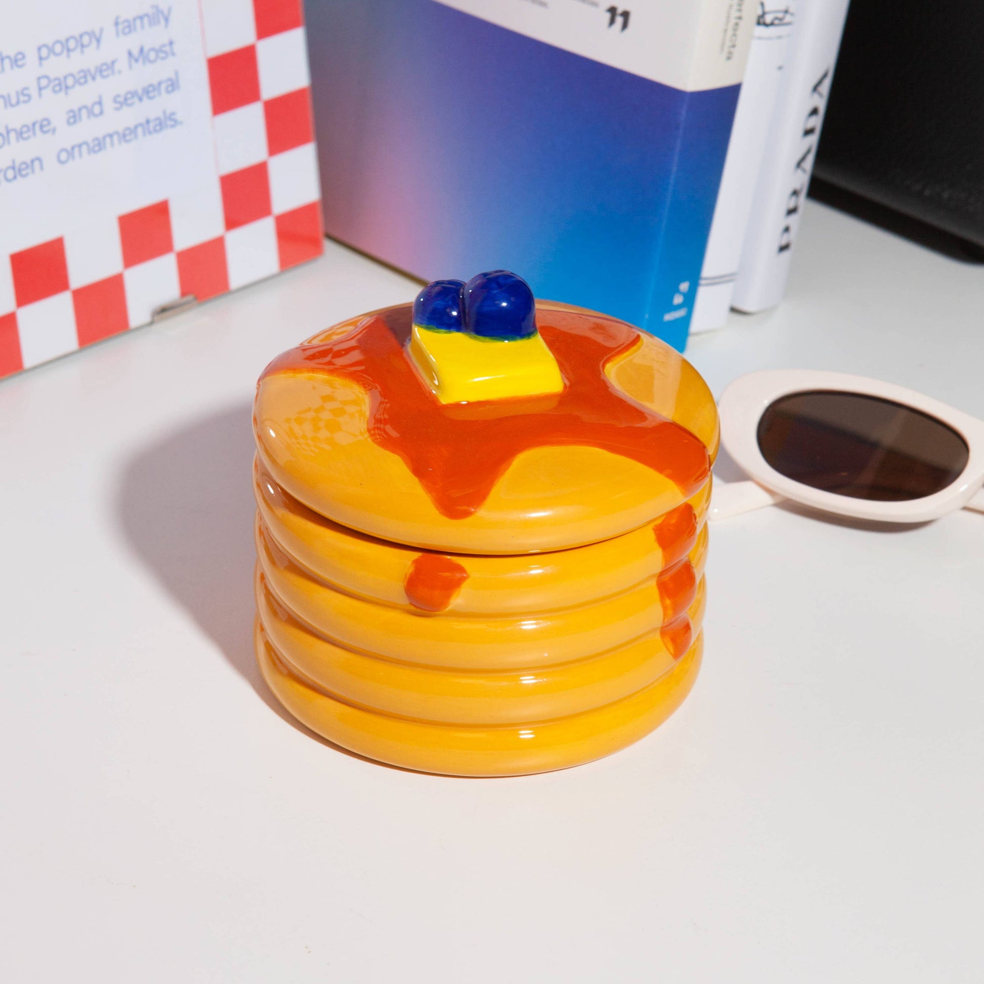 Ceramic storage jar made to look like a stack of pancakes with a decorative lid with a pad of butter and blueberries on a white background with books in background 