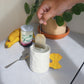 white, ceramic mug with a yellow handle shaped like farfalle pasta with water inside and a tea bag about to be dropped in 