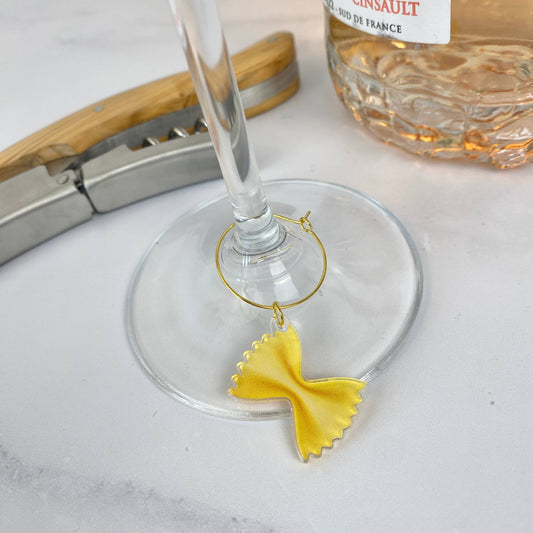 Wine glass with a yellow bow tie pasta drink charm on a white surface, with a bottle of wine in the background.