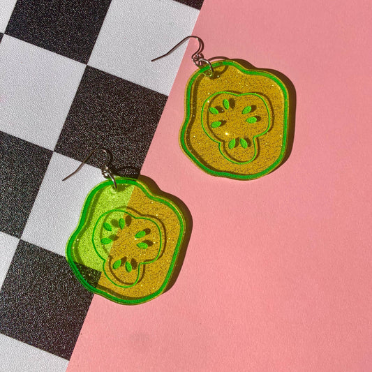 Glittery, green, acrylic, sliced pickle earrings on fish hook backings on a half pink and black & white checkerboard background 