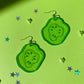 Glittery, green, acrylic, sliced pickle earrings on fish hook backings on green background with mini star confetti pieces 