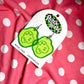 Glittery, green, acrylic, sliced pickle earrings on fish hook backings with cardboard backing/packaging. Photographed on pink and white polka dot background