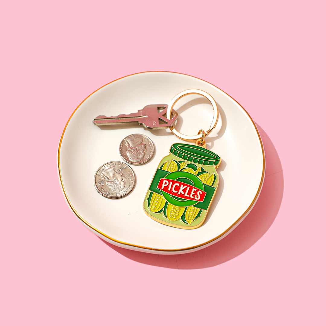 Keychain with pickle jar design on a white dish with coins and keys against a pink background
