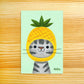 A square art print featuring a cute gray tabby cat wearing a yellow pineapple costume, displayed flat on a light wood grain background. The print has a pale mint green background and is signed 'Nellie' in the bottom right corner.