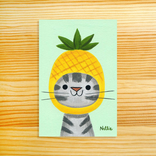 A square art print featuring a cute gray tabby cat wearing a yellow pineapple costume, displayed flat on a light wood grain background. The print has a pale mint green background and is signed 'Nellie' in the bottom right corner.