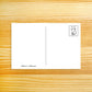 A white postcard with a vertical center line, a small black cat illustration in the top-right corner, and the text 'Nellie Le - nellilele.com' at the bottom left, placed on a light wood grain background.