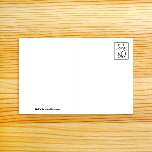 A white postcard with a vertical center line, a small black cat illustration in the top-right corner, and the text 'Nellie Le - nellilele.com' at the bottom left, placed on a light wood grain background.
