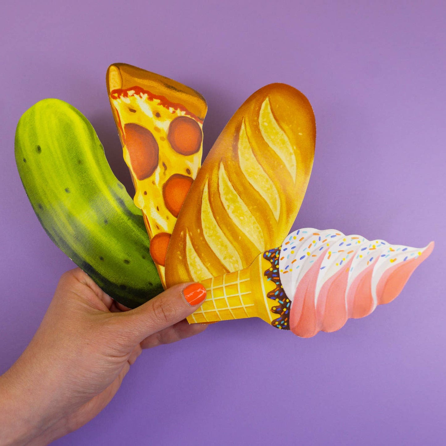 Hand holding bookmarks shaped like a cucumber, pizza slice, baguette, and ice cream cone against a purple background