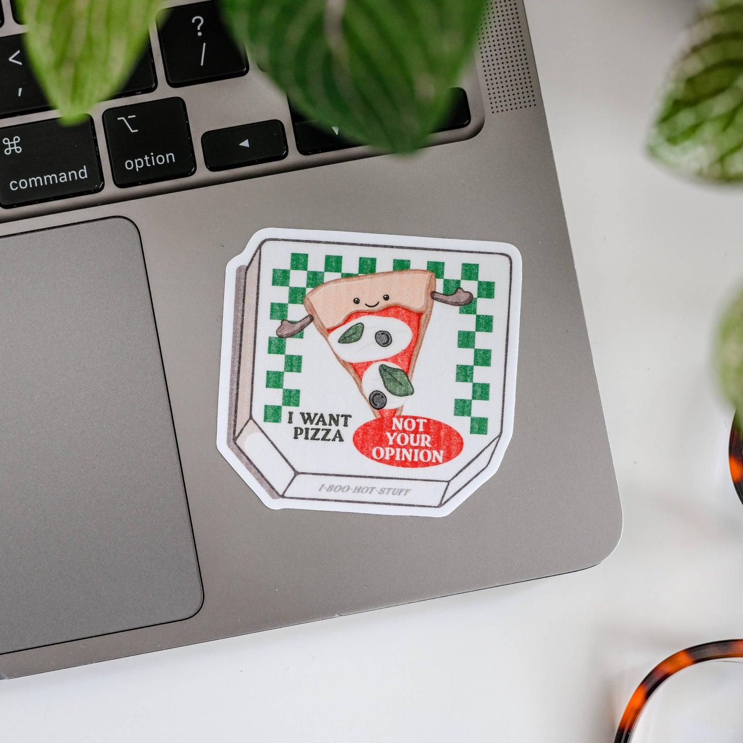 Laptop with a 'I Want Pizza Not Your Opinion' sticker on a white surface with glasses and plants.