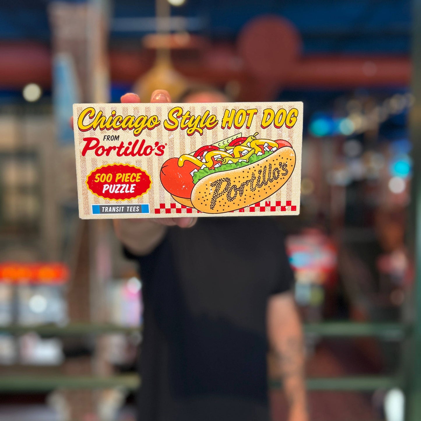 Person holding a Portillo's hot dog puzzle box in front of a blurred indoor background