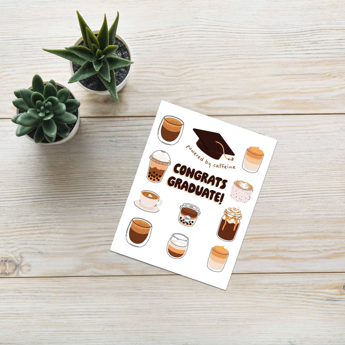 Greeting card with coffee-themed designs and 'Congrats Graduate!' text on a wooden surface with plants.