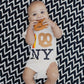 Baby wearing a onesie with 'NY' and pretzel design, sitting on a black and white zigzag patterned blanket with the pretzel teether in it's mouth.