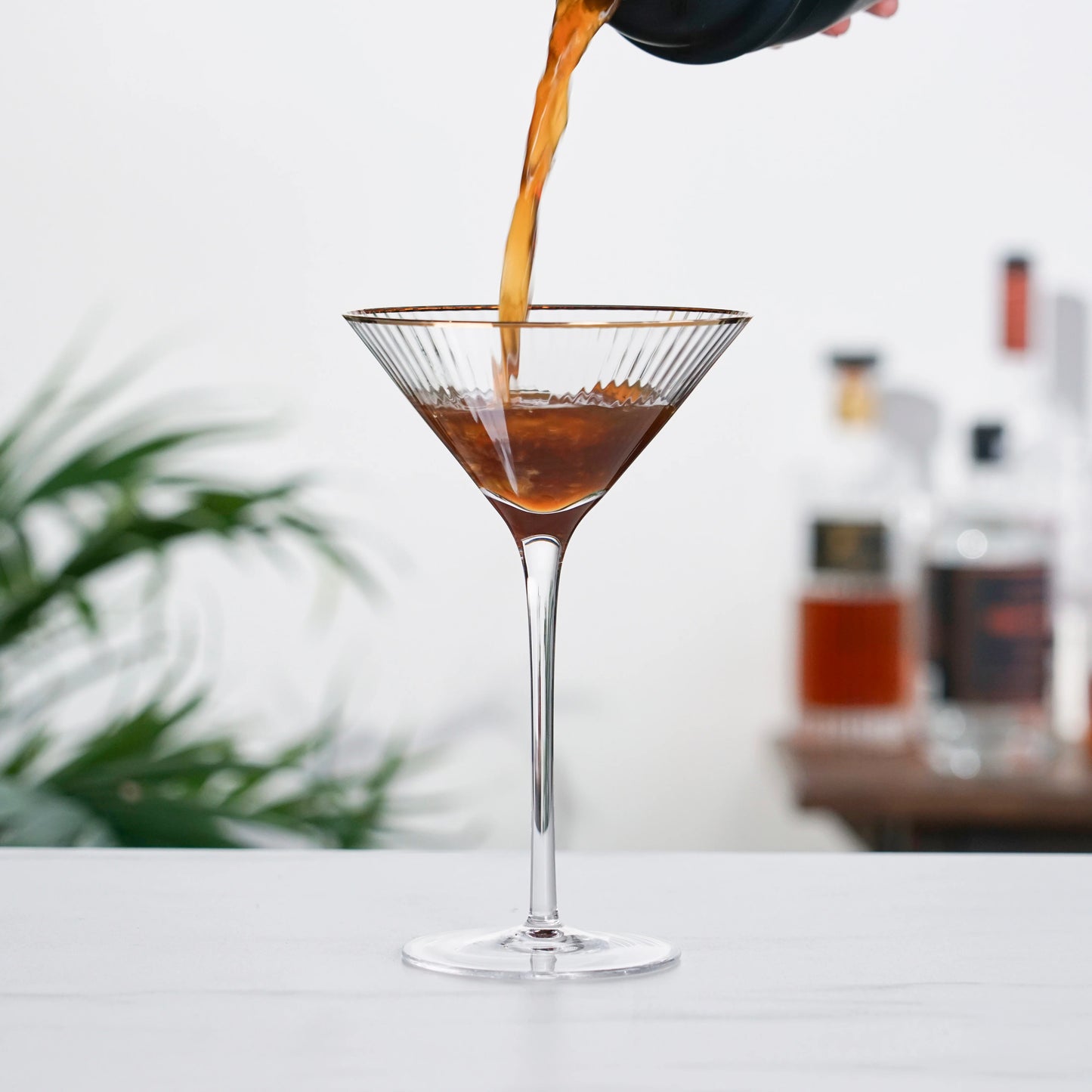 Photo of a dark brown cocktail being poured from a shaker into a single rippled crystal martini glass with gold dipped rim.