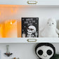 A styled shelf display featuring a black greeting card with a white skeleton graphic and the text 'YOU'RE RAD TO THE BONE' next to a white plush ghost toy. The shelves are white with gold handles, flanked by glowing orange candles, a small potted plant, and other Halloween-themed decor.