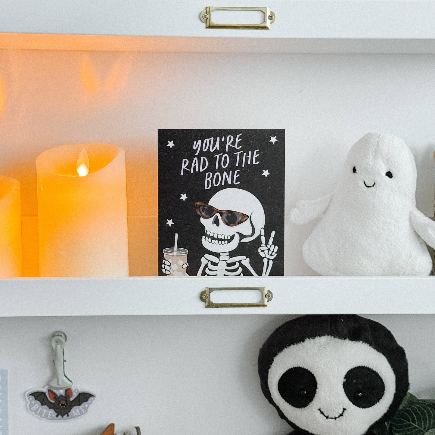 A styled shelf display featuring a black greeting card with a white skeleton graphic and the text 'YOU'RE RAD TO THE BONE' next to a white plush ghost toy. The shelves are white with gold handles, flanked by glowing orange candles, a small potted plant, and other Halloween-themed decor.