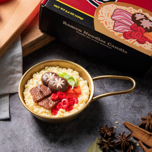 Candle shaped like a pot of ramen with ingredients on a dark surface, next to its packaging.