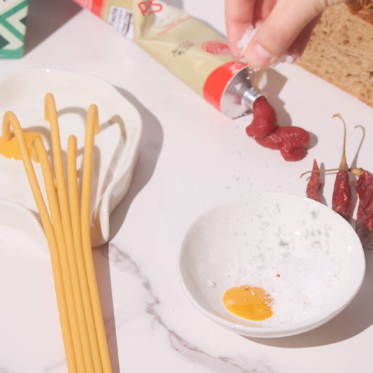 Hand holding a pinch of salt over a tube of tomato paste, next to the egg spoon rest and egg bowl, surrounded by ingredients on a light surface.