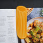 Pasta-shaped bookmark on an open cookbook with pasta dish in the background