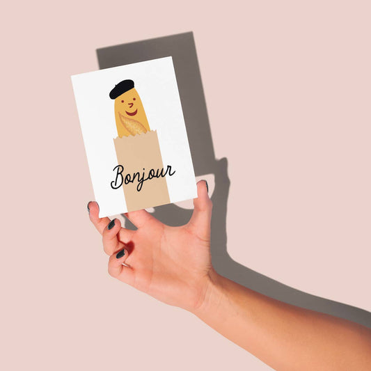 Greeting card with a smiley baguette wearing a beret on it. Text reads "bonjour" -- card is being held by a hand against a wall 