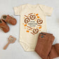 Flat lay of a baby onesie that reads "more snacks please" in the center of various snacks including cheerios, pretzels and goldfish, along with brown baby pants, shoes and a wooden toy