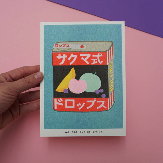 Hand holding a risograph print with a retro-style product illustration of Sakuma Drops, against a pink and purple background