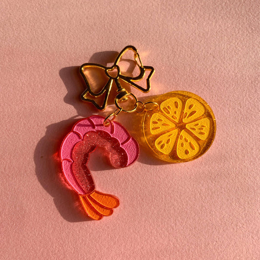 Three colorful charms, shrimp, lemon and bow on a pink background