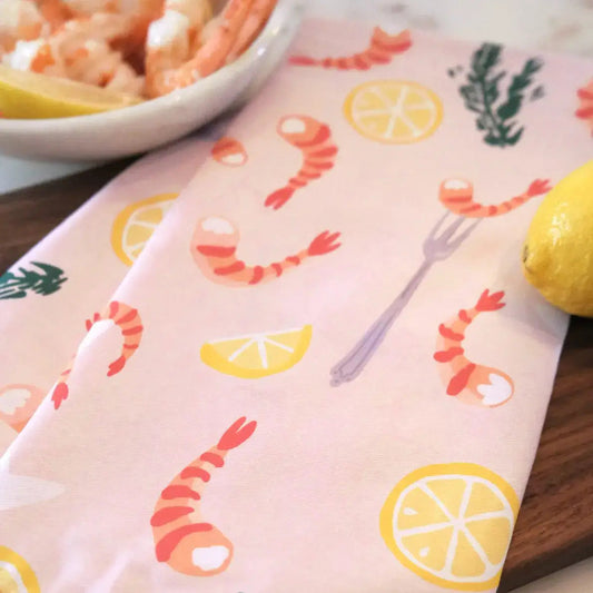 Kitchen tea towel, light pink/salmon in color with a design of illustrated lemon halves and wedges, shrimp, cocktail fork and herbs