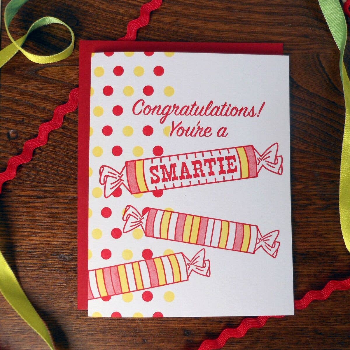 Greeting card with Smarties candy design and 'Congratulations! You're a SMARTIE' text on a wooden surface with ribbons.