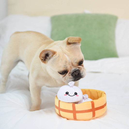 Dog playing with a plush soup dumpling dog toy in a steamer basket on a bed