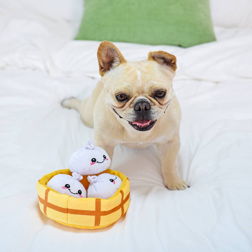 Dog sitting on a bed with a plush toy featuring 3 soup dumplings in a steamer basket 