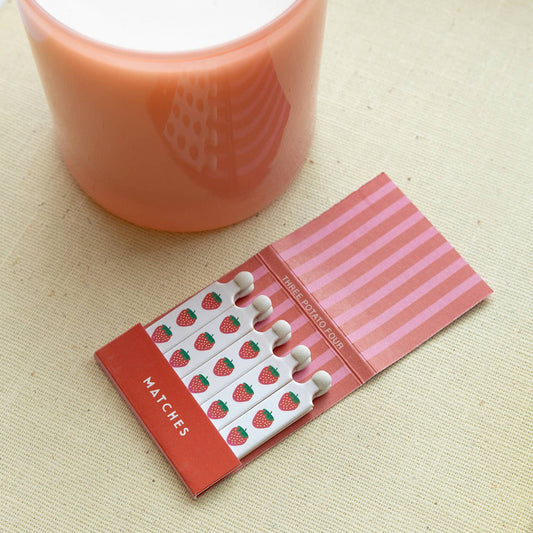 Matchbook with strawberry design next to a pink candle on a beige surface