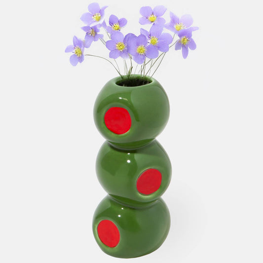 Vase shaped like 3 stuffed green olives stacked on top of each other containing purple flowers on a white background