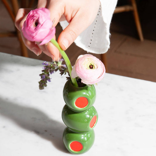 Stuffed green olive vase holding pink flowers on a white surface